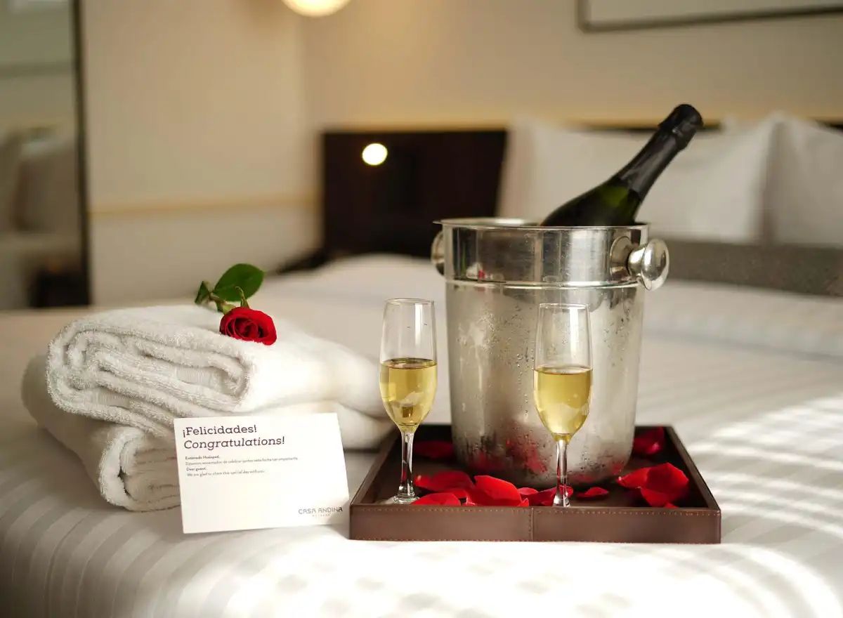 Champagne bottle with two glasses, rose petals, folded towels, and a celebration card arranged on a hotel bed.