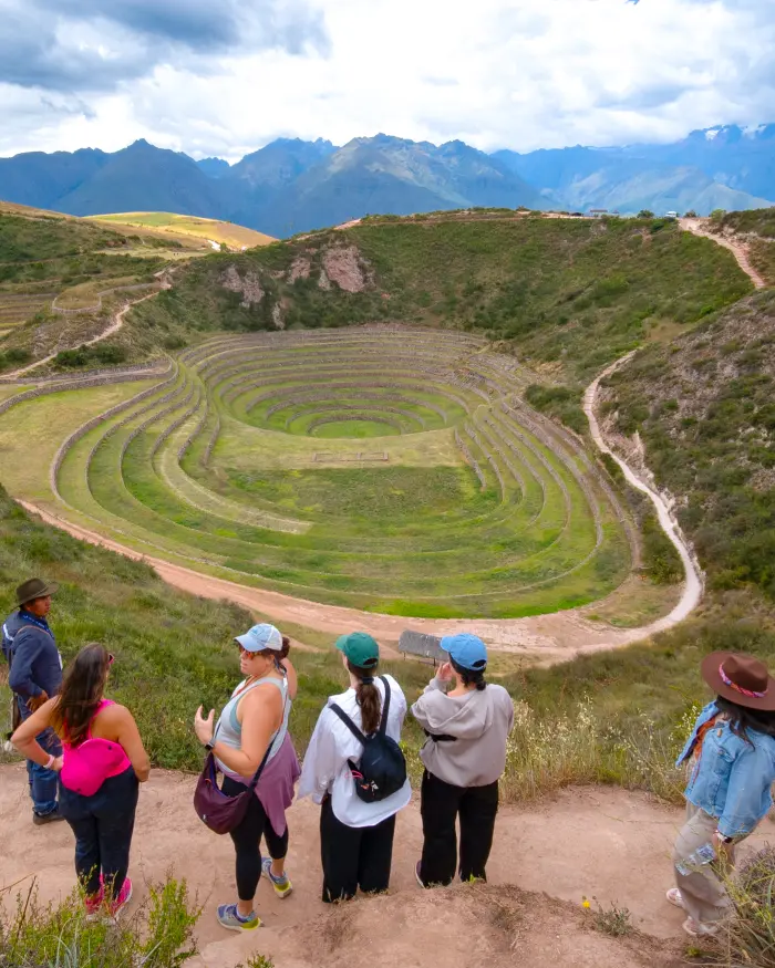 Centro arqueológico de Moray en Cusco con terrazas circulares, turistas observando el paisaje y montañas andinas de fondo.