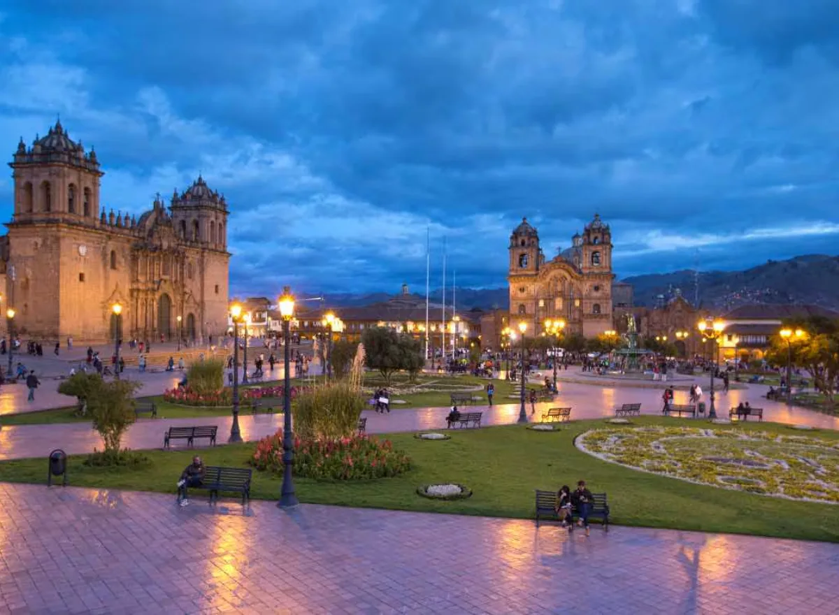 Cusco Historic Center at night with illuminated Plaza de Armas, cathedrals, gardens, benches, and a dark blue sky.