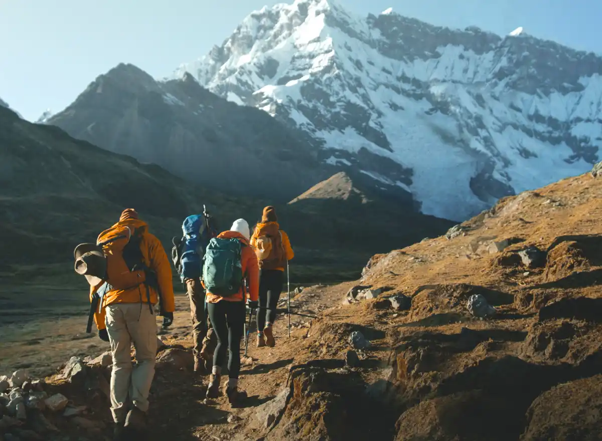 Travelers on a multi-day trek in the Andes with backpacks, a rocky trail, mountain scenery, and a snow-covered peak in the background.