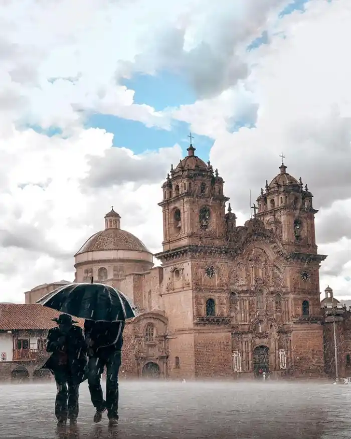 Travelers walking with an umbrella in the rain at Cusco's Plaza de Armas, in front of the Cathedral under a cloudy sky.