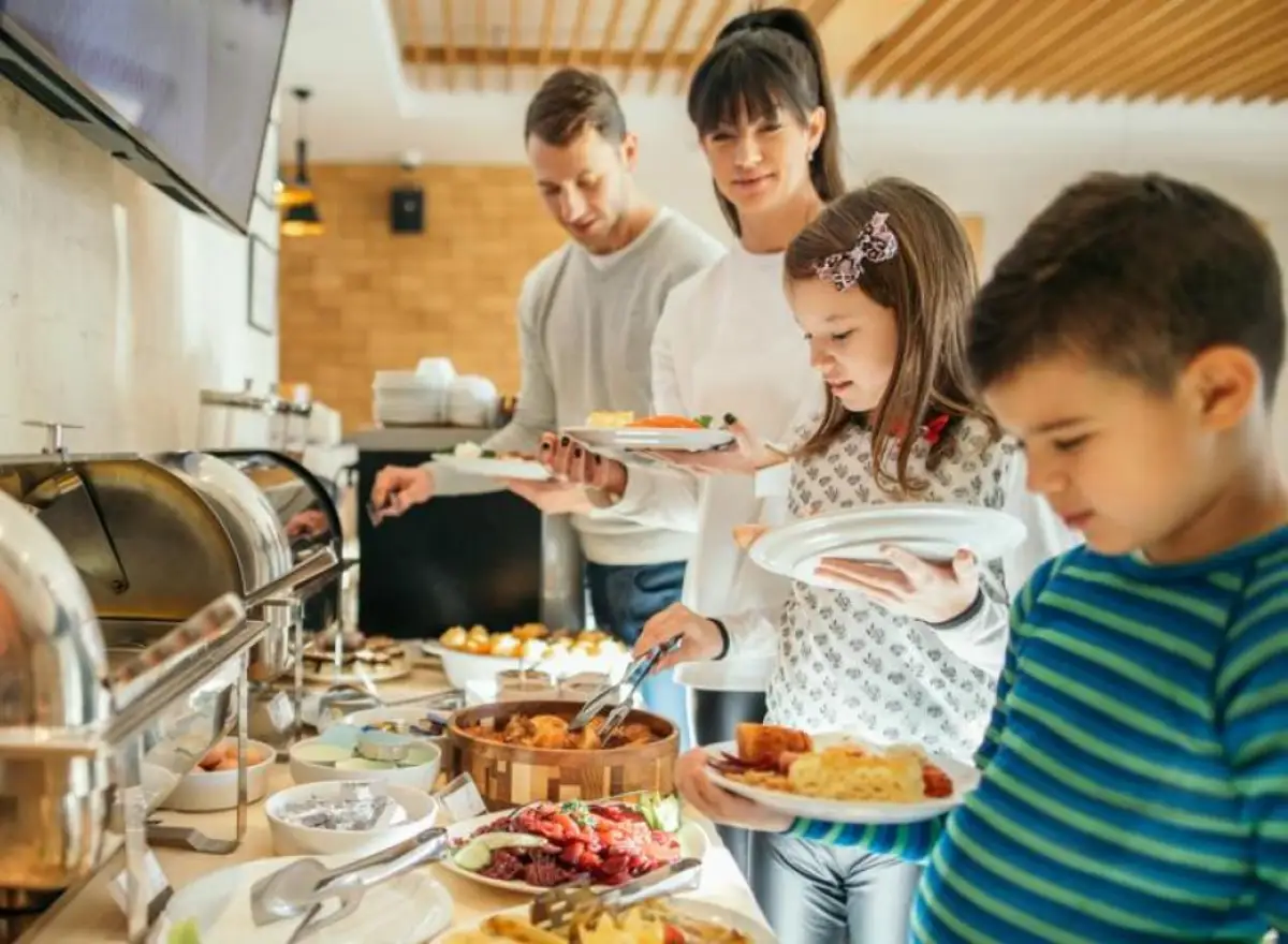 Familia sirviéndose desayuno buffet en un hotel, con niños, platos variados y alimentos calientes antes de iniciar el día.