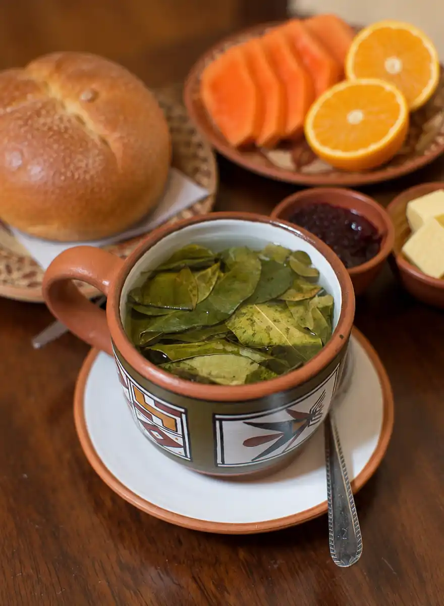 Coca tea served in an Andean-style cup with chuta bread, fruit, cheese, jam, corn, and other Cusco breakfast items.