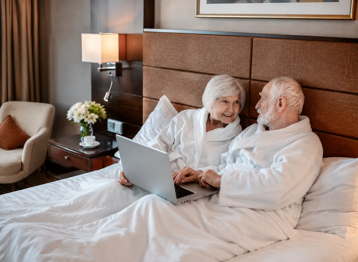 Older couple resting in a hotel room on the bed with a laptop, white robes, large windows, and a comfortable atmosphere.