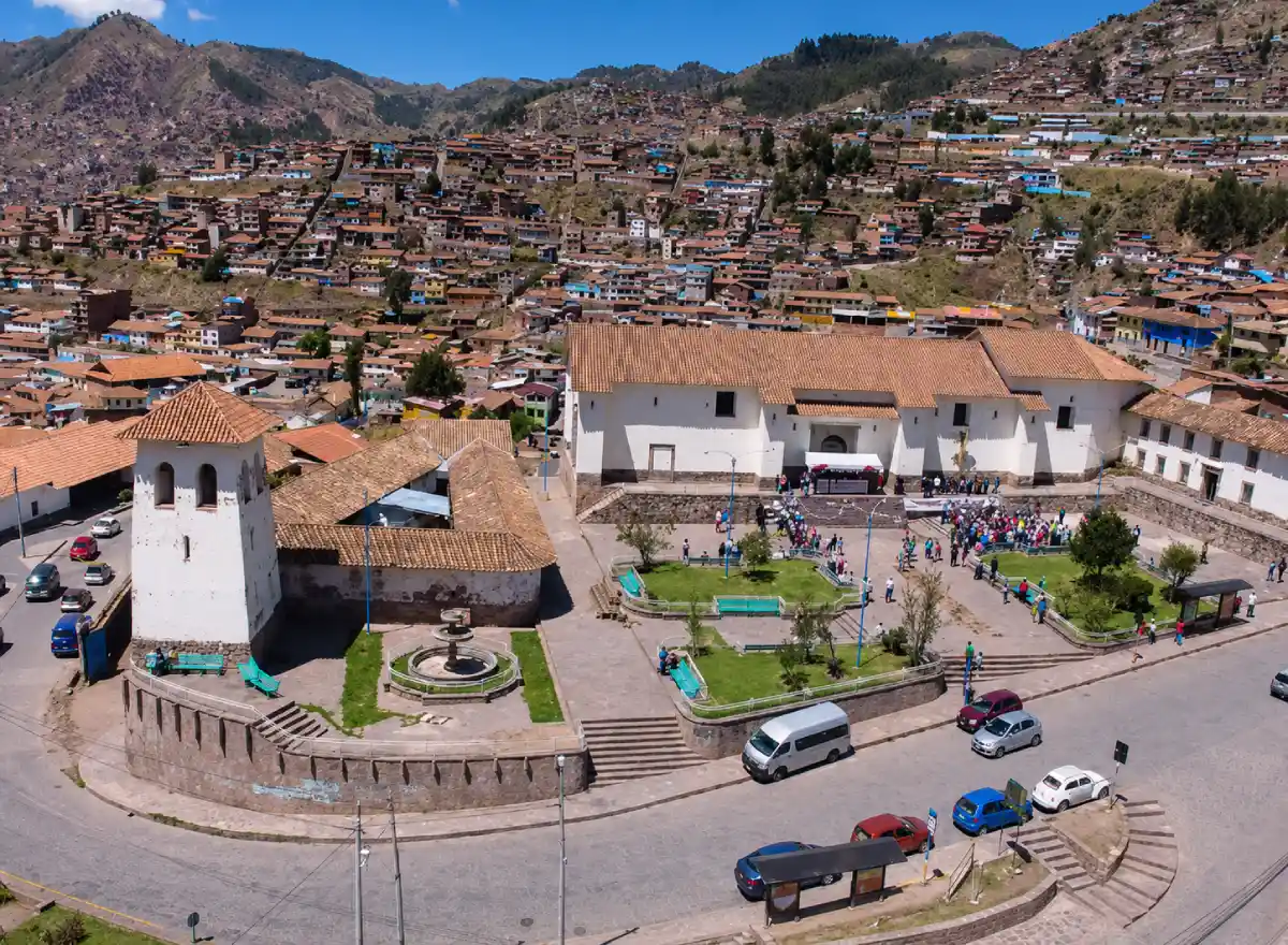Santa Ana square in Cusco with a church, green areas, vehicles, visitors, and city views from an elevated neighborhood.