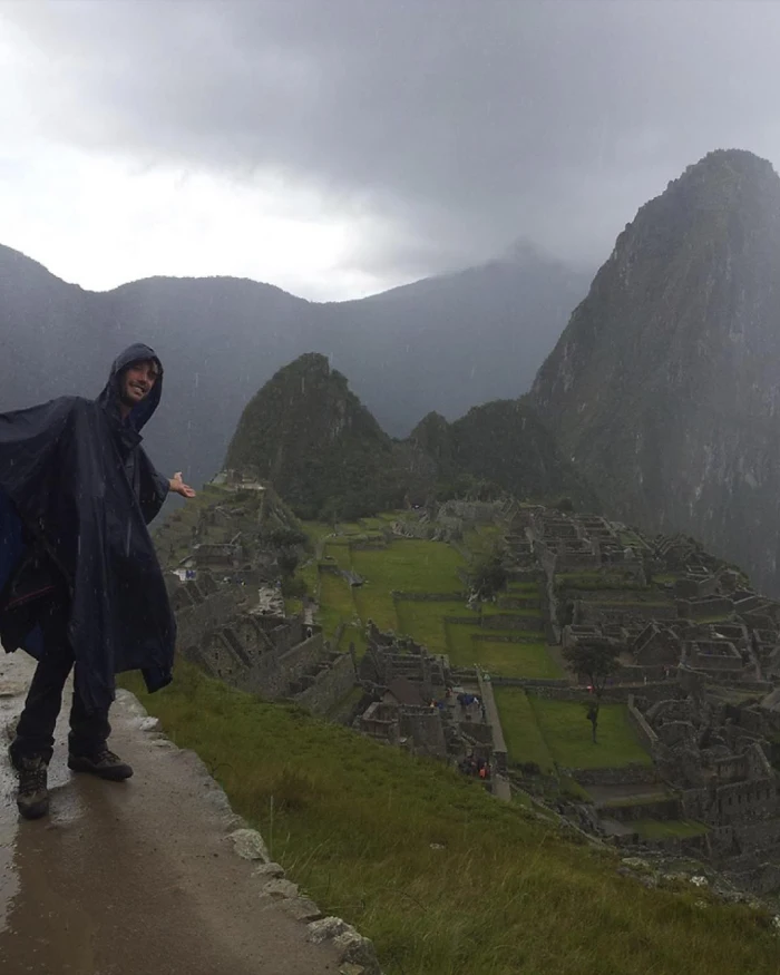 Traveler wearing a waterproof poncho at Machu Picchu during the rainy season, with the citadel and cloudy mountains in the background.